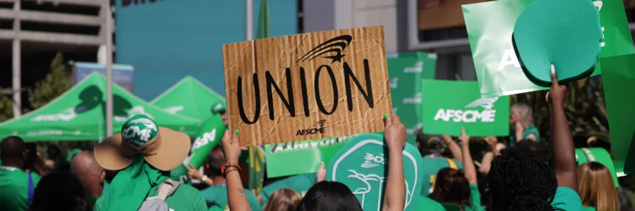 AFSCME members in green shirts at a rally. In the center, one holds a "union" sign.